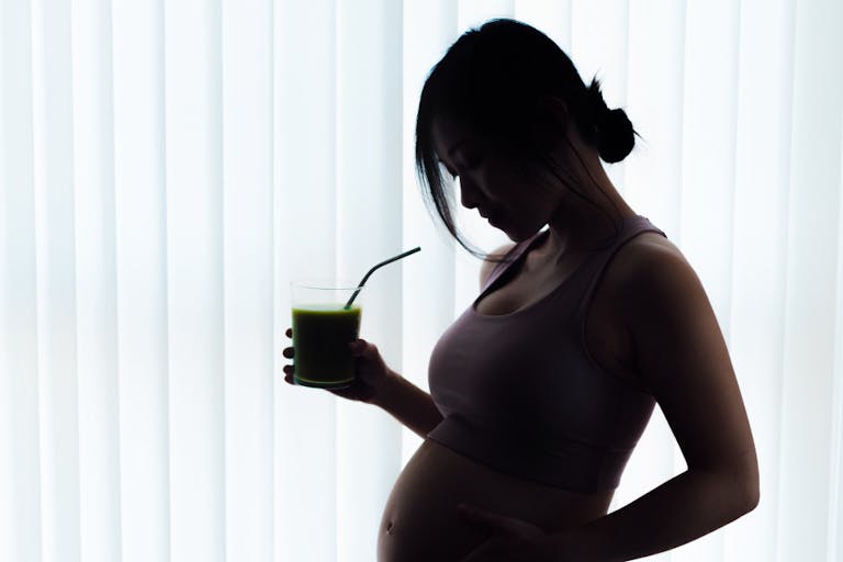 Side view of silhouette pregnant woman holding a glass of drink and standing by window at home