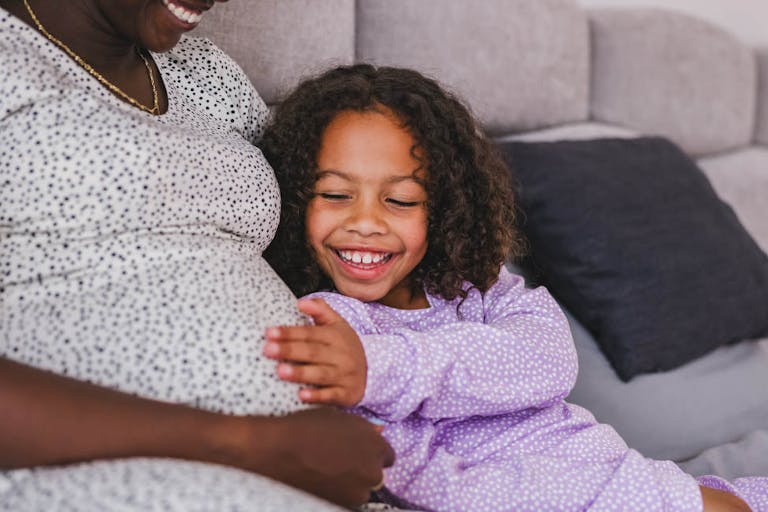 Photo: AzmanJaka/Getty Images Happy Black Little Daughter Touching Her Black Pregnant Mother’s Belly While They Are Both Lying In A Bed In Pajamas
