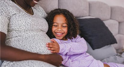 Happy Black Little Daughter Touching Her Black Pregnant Mother’s Belly While They Are Both Lying In A Bed In Pajamas