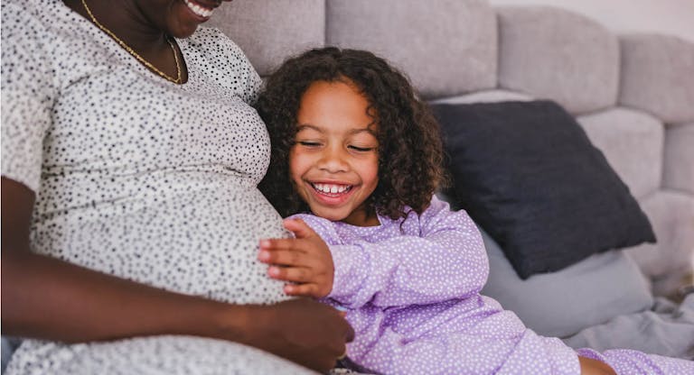 Happy Black Little Daughter Touching Her Black Pregnant Mother’s Belly While They Are Both Lying In A Bed In Pajamas