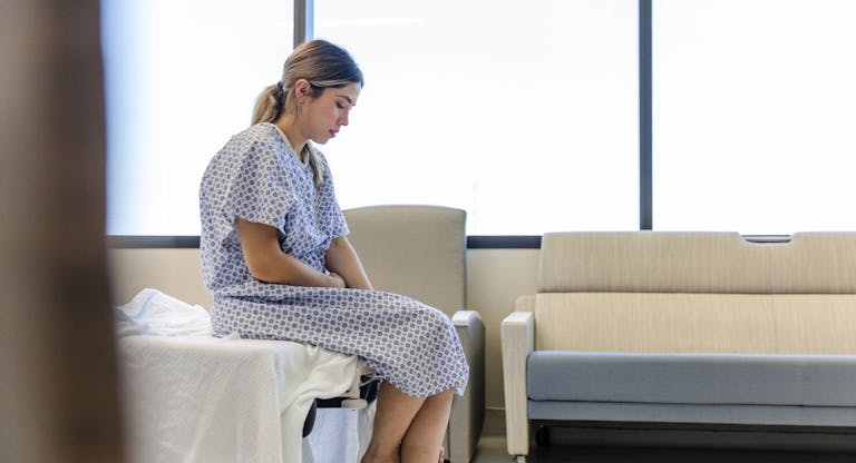 Anxious, sad, young woman wearing hospital gown looks down
