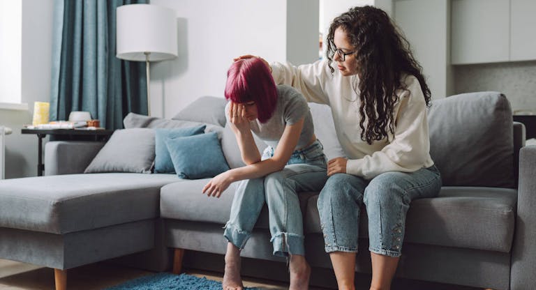 Adult curly haired woman consoling, stroking teen girl, sitting on a sofa. (Getty Images)