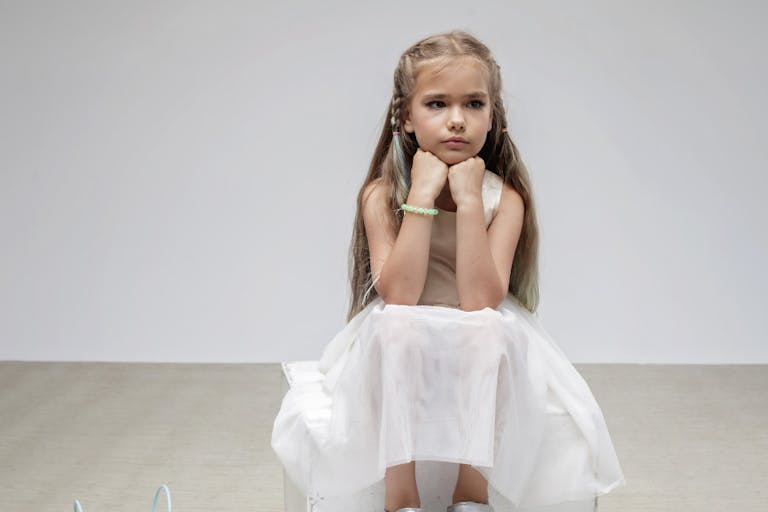 Sad girl with long hair in festive dress sits on square podium, white background, studio shot