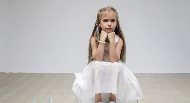 Sad girl with long hair in festive dress sits on square podium, white background, studio shot