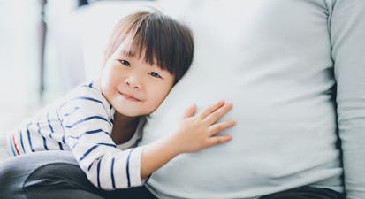 Cute little Asian girl touching and listening to pregnant mother’s belly with joy