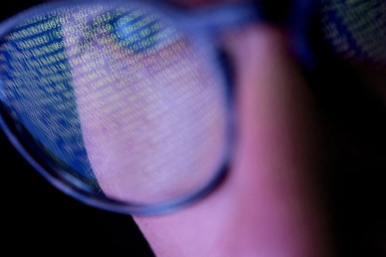 A person with glasses in a dark room looking at computer screen that’s displaying codes or programming, with reflections on the glasses keeping the subject engrossed in