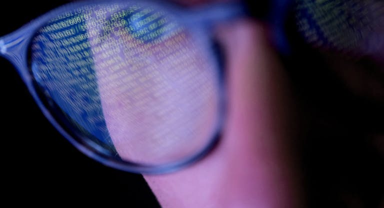 A person with glasses in a dark room looking at computer screen that’s displaying codes or programming, with reflections on the glasses keeping the subject engrossed in