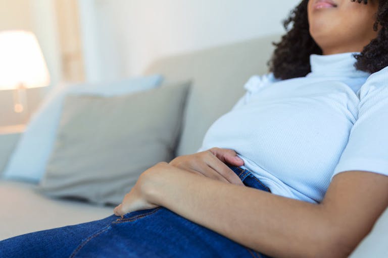 African American Woman in painful expression holding hands against belly suffering menstrual period pain, lying sad on home bed, having tummy cramp in female health concept