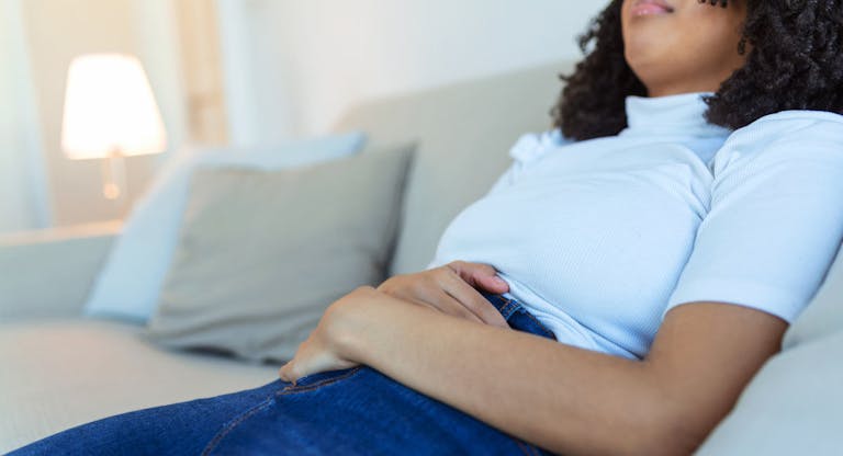 African American Woman in painful expression holding hands against belly suffering menstrual period pain, lying sad on home bed, having tummy cramp in female health concept