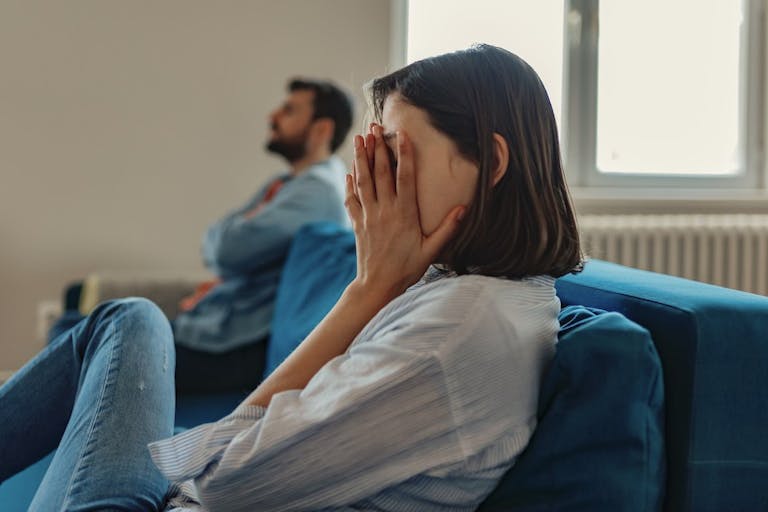Photo: Getty Images A woman sits on a couch with her hands covering her face. In the background, a man sits crossing his arms.