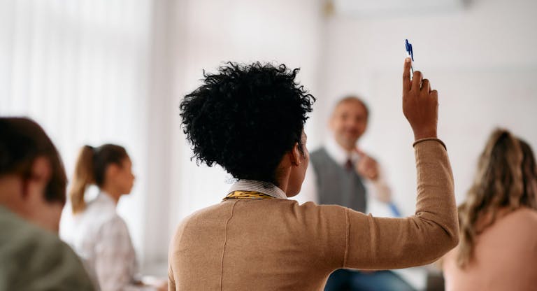 Rear view of black student raising her hand to answer a question during a class at lecture hall.