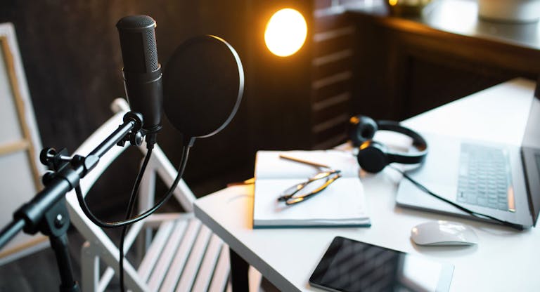 Podcast streaming at home. Audio studio with laptop, microphone with pop filter and headphones on white table against black wall with warm lights. Blogger concept.