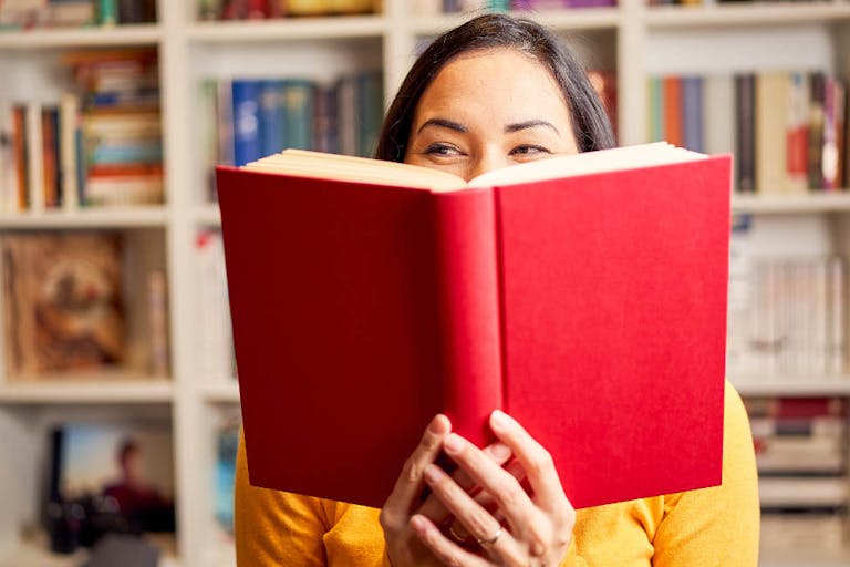 Female young behind book with face covered for a red book while smiling