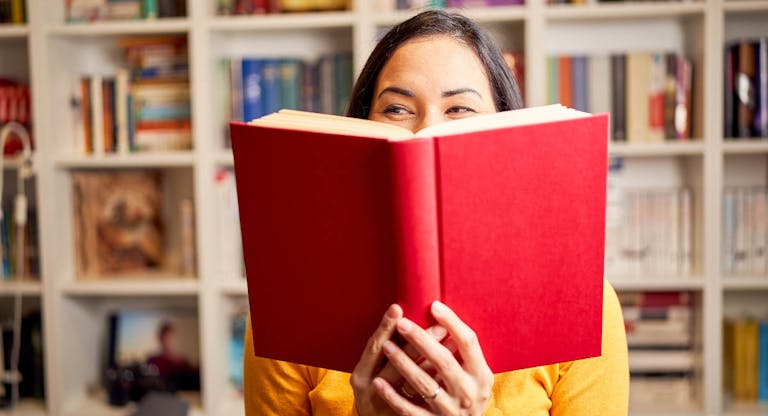 Female young behind book with face covered for a red book while smiling