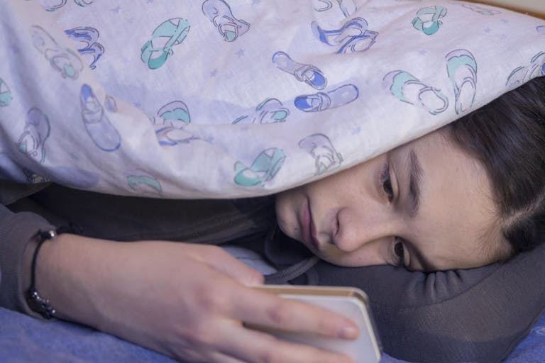 Girl using phone in bed While Lying Under Blanket during the day, Front view, copy space Photo of Girl using phone in bed during the day