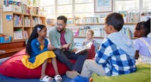 Male teacher reading a book to students while sitting in school library. Librarian read a fairy tale to a group of multiethnic primary schoolchildren. Middle eastern man at elementary school reading stories to little boys and curious girls.