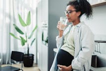 A pregnant woman enjoys a refreshing glass of water in a contemporary kitchen. The image captures a serene and healthy lifestyle moment, highlighting the importance of hydration during pregnancy. Pregnant Woman Drinking Water in Modern Kitchen Setting