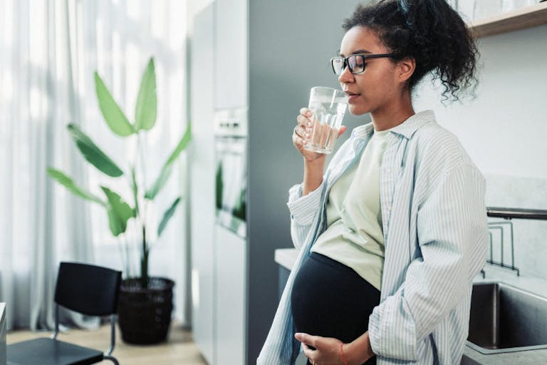 A pregnant woman enjoys a refreshing glass of water in a contemporary kitchen. The image captures a serene and healthy lifestyle moment, highlighting the importance of hydration during pregnancy. Pregnant Woman Drinking Water in Modern Kitchen Setting