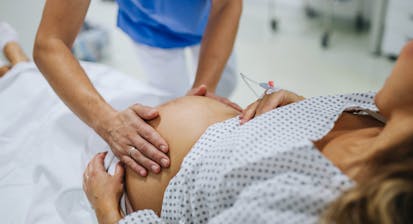 Obstetrician examining pregnant woman in hospital, touching woman’s belly, examine position of baby, breach position, size of baby and movements. Woman in labour laying on hospital bed, talking with ob-gyn doctor in maternity ward.