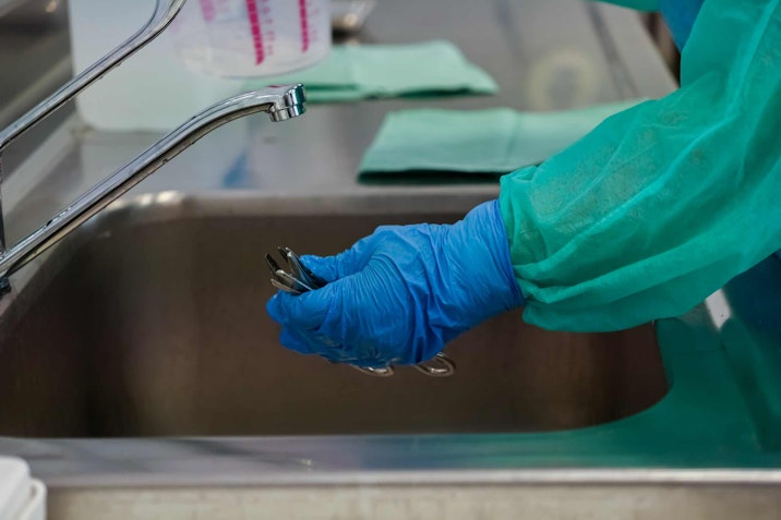 A person in blue protective gloves and a green gown carefully washes or cleans medical instruments in a sterile stainless steel sink, focusing on hygiene and sanitation procedures medical staff at faucet with rubber gloves