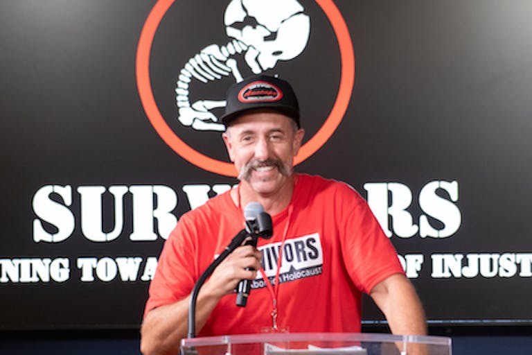 Photo courtesy of Tim Clement, Director, Survivors of the Abortion Holocaust Tim Clement stands before a banner for the Survivors of the Abortion Holocaust while holding a microphone.