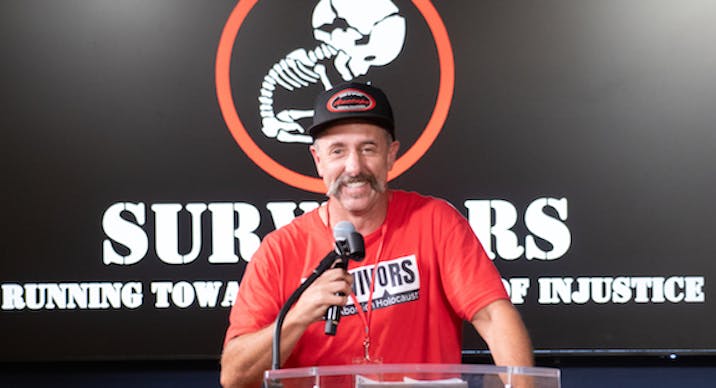 Tim Clement stands before a banner for the Survivors of the Abortion Holocaust while holding a microphone.