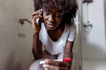 A woman talking on the phone with her while sitting on the toilet bowl and holding a home pregnancy test.