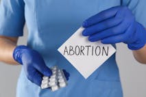 Doctor holding pills and paper card with word ABORTION on light grey background, closeup