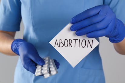 Doctor holding pills and paper card with word ABORTION on light grey background, closeup