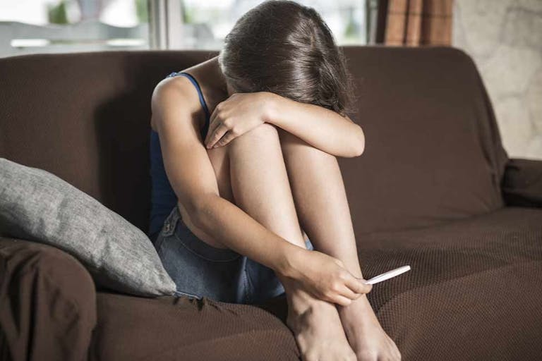 teen girl sitting on couch with her head in her hands, holding a pregnancy test