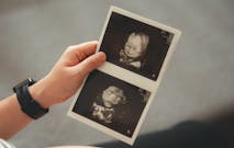 Close-up shot of pregnant woman holding and looking at ultrasound scans, sitting on sofa in living room at home.