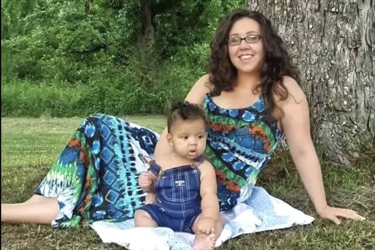 Mother sits by a tree with her young son, who is dressed in bib overalls
