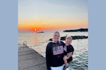 Gabby Weiss stands on a dock holding her young son with an orange sunset behind them.