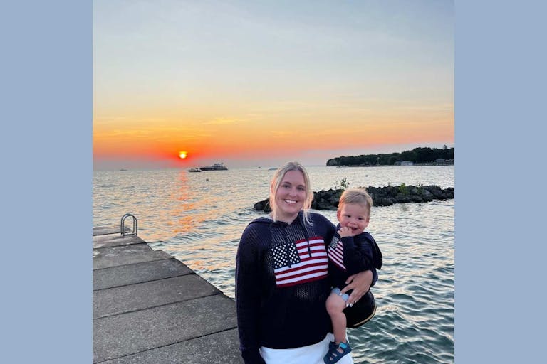 Gabby Weiss stands on a dock holding her young son with an orange sunset behind them.