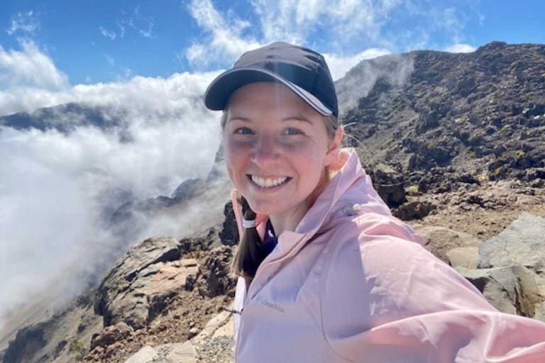 Jane Allen. Photo courtesy of her family. Jane Allen smiles as she stands on top of a rocky mountain wearing a pink jacket and gray baseball hat. A blue sky with white clouds around her.