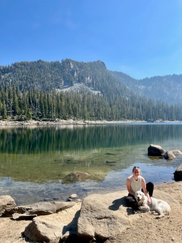 Jane Allen at a lake with her dog. Photo courtesy of the Allen family. Jane Allen and her dog sit on a rock next to lake with evergreen trees across the water.