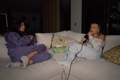 Image shows two women sitting on a couch in their pajamas with a box of tissues sitting between them.