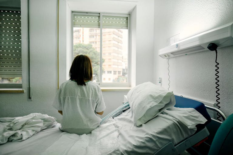 Photo: Yoss Sabalet/Getty Images sick woman alone sitting on the bed of a hospital room looking out the window seen from behind
