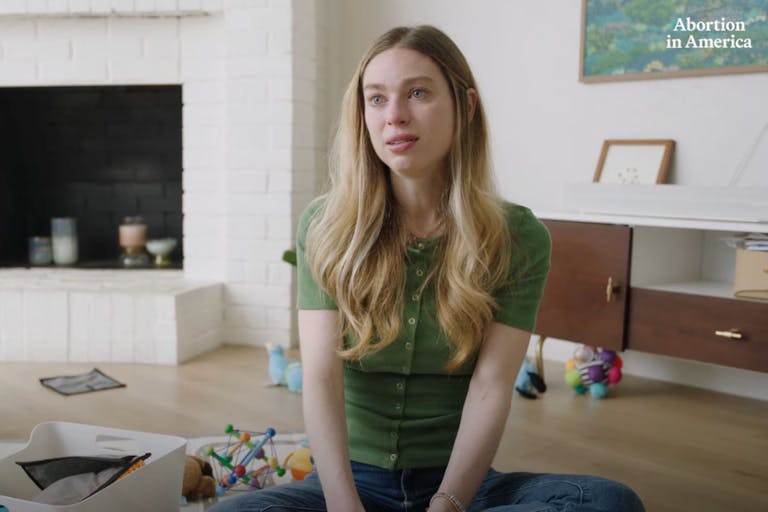 Screenshot: 'Abortion in America' (YouTube) Woman with long blonde hair and a green shirt sits on the floor in a living room surrounded by toys.