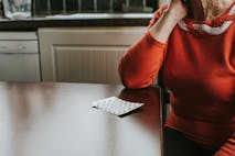 A blister pack of tablets sits on a wooden table. An elderly woman rests her chin on her hand and looks at the medication infront of her.