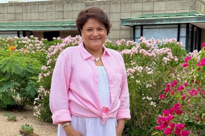 Image shows a woman with short brown hair and a pink blouse, smiling in front of a garden.