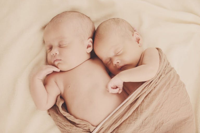 Image shows two babies snuggled in a tan blanket.