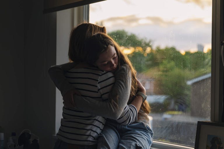 Photo: Justin Paget/Getty Images Powerful view of a young teenage girl being comforted by her mother as she sits on her window sill at sunset