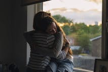 Powerful view of a young teenage girl being comforted by her mother as she sits on her window sill at sunset