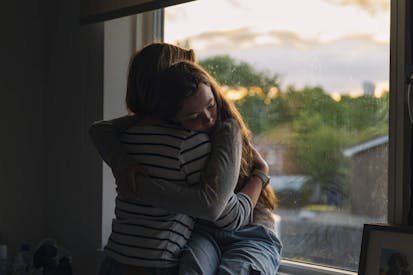 Powerful view of a young teenage girl being comforted by her mother as she sits on her window sill at sunset