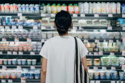 Rear view of young Asian woman doing grocery shopping, standing in front of a shelf full of fresh dairy products in the dairy aisle and thinking which product to choose from in a supermarket. Concept of customer awareness
