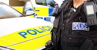 Close-up of anonymous police officer wearing stab proof vest uniform and gear worn by constablury at the scene of an event. They are in front of emergency services police force vehicles featuring a cop car and police van, in response to an incident. Officers provide law enforcement, establish public order and protect citizens.