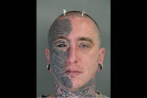 Photo shows a mugshot of a man with tattoos covering half his face, metal spikes protruding from the top of his head, lip piercings, and gauged ears.