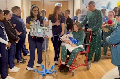 Image shows mom in a hospital wheelchair carrying her baby as a nurse leads them through a hallway lined with hospital staff. The baby's father pushes the wheelchair.