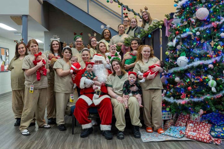 Photo: Missouri Department of Corrections/Facebook A group of female inmates in beige jumpsuits hold their babies next to a large Christmas tree, with Santa Claus in the middle.